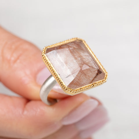 Ring with a gold and silver band and a rutilated quartz stone held by a hand on a light background