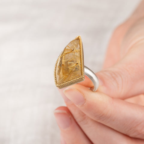 A hand holding a rutilated quartz ring with an 18k gold bezel and sterling silver band