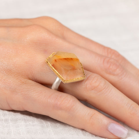 Hand wearing a ring with a large, faceted rutilated quartz gemstone on a light background
