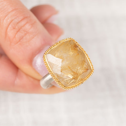 Gold and silver ring with a rutilated quartz gemstone held by a hand on a light background