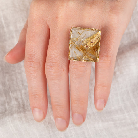 A hand wearing a handcrafted ring made with a rutilated quartz, 18k gold bezel and sterling silver band