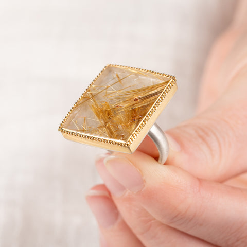 A close up of a hand holding a handcrafted ring made with a rutilated quartz, 18k gold bezel and sterling silver band