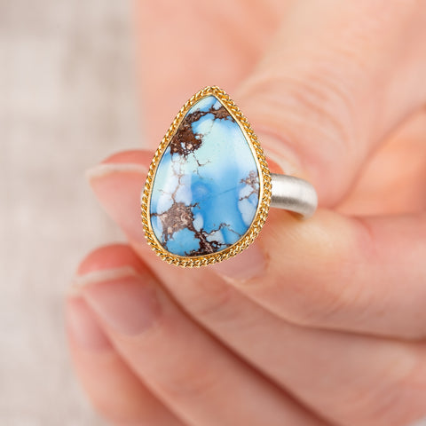 Close up of a hand holding a turquoise teardrop ring with gold bezel and sterling silver band