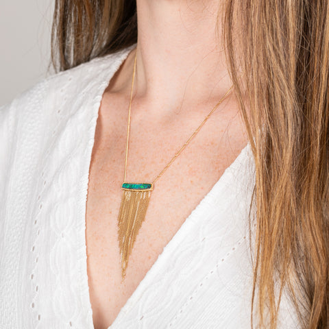 A person wears a handcrafted necklace featuring a boulder opal gemstone and 18k gold tassel adornment
