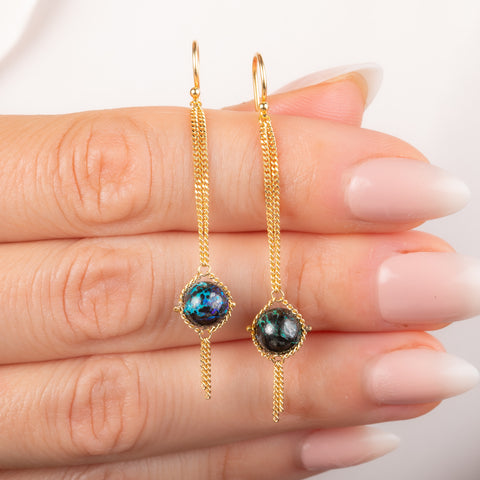 A pair of gold dangle earrings with round iridescent blue and green boulder opal stones, shown hanging from a person's fingers against a clean white background.