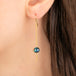 A close-up of a gold dangle earring worn on a person's ear, featuring a round iridescent boulder opal stone with a long gold chain