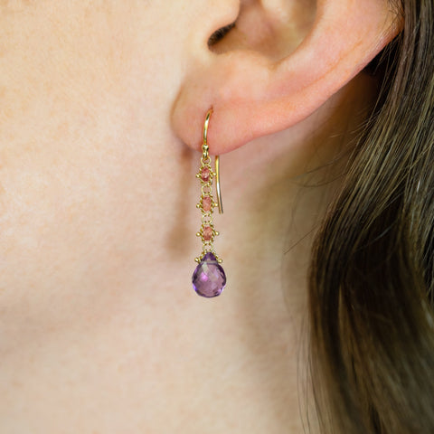 A close-up of an 18k yellow gold drop earring that features three woven spinel beads and a teardrop shaped amethyst hanging from a french hook closure