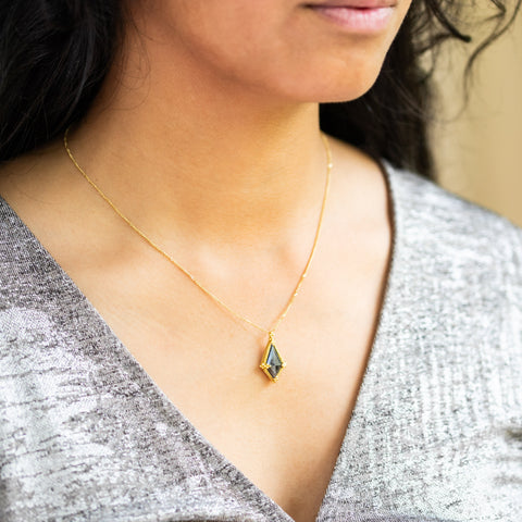 A model wears a Kite-shaped Black Diamond pendant which is set in an 18k yellow gold bezel, suspended from an 18K yellow gold chain. Set in a distinctive one-of-a-kind design with handmade gold bezel, braided detail, and granulation.