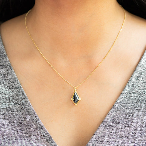 A model wears a Kite-shaped Black Diamond pendant which is set in an 18k yellow gold bezel, suspended from an 18K yellow gold chain. Set in a distinctive one-of-a-kind design with handmade gold bezel, braided detail, and granulation.