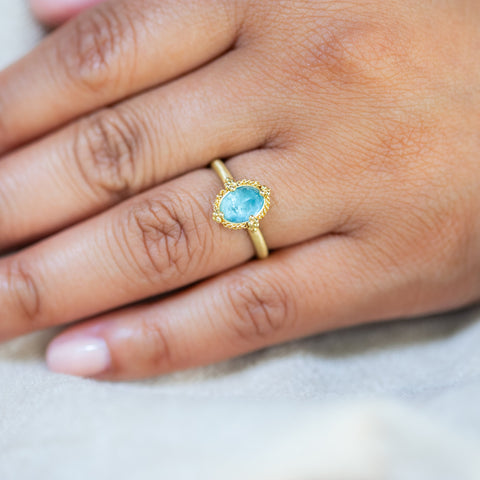 A model wears this Aquamarine ring in 18K yellow gold, featuring a misty blue oval gemstone reflecting a serene summer sky. Meticulously handmade gold bezel with braided gold accents and granulated prongs. Handmade in New York
