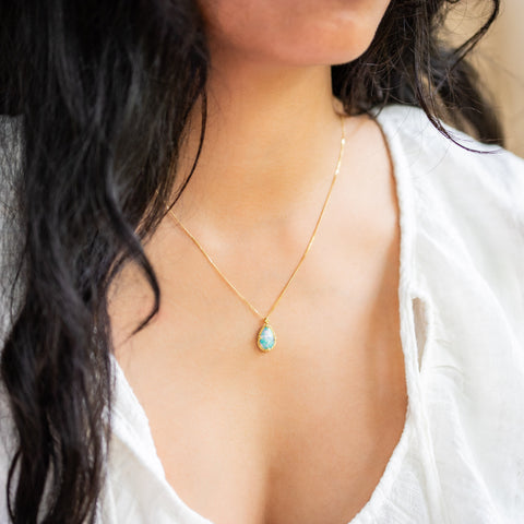 A model wears an 18K yellow gold Ethiopian Opal pendant. Iridescent colors include green, blue, and pink. Suspended from a handmade frame with braided accents on an 18K yellow gold chain. Handmade in New York.