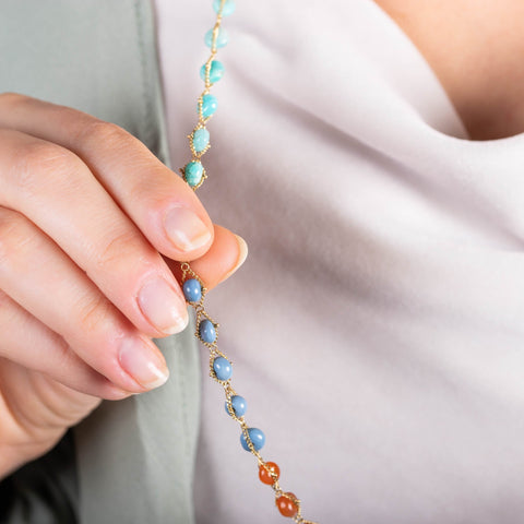 A close up of a model holding a woven necklace with amazonite, sunstone, and blue and pink opal beads threaded with 18k yellow gold. 
