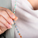 A close up of a model holding a woven necklace with amazonite, sunstone, and blue and pink opal beads threaded with 18k yellow gold. 