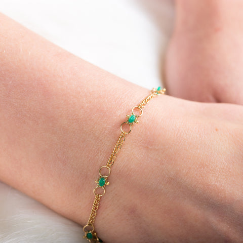 A model wears a delicate 18k yellow gold chain bracelet that is dotted with emerald beads throughout. The bracelet is finished with a lobster clasp closure.