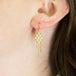 A model wears an 18k yellow gold earring crafted with white pearls woven into a diamond shaped lattice pattern and has three dangling chains at the bottom.