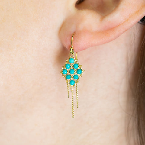 A model wears an 18k yellow gold earring crafted with turquoise beads woven into a diamond shaped lattice pattern and has three dangling chains at the bottom.
