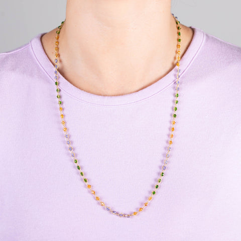 A model wears a long woven chain necklace that features Green Tourmaline, Garnet, and Tanzanite stone beads woven in 18k yellow gold chain.