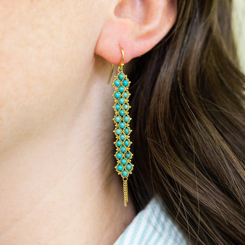 A close up of a pair of long turquoise earrings which feature a woven lattice pattern and two dangling chains at the bottom of the earring.