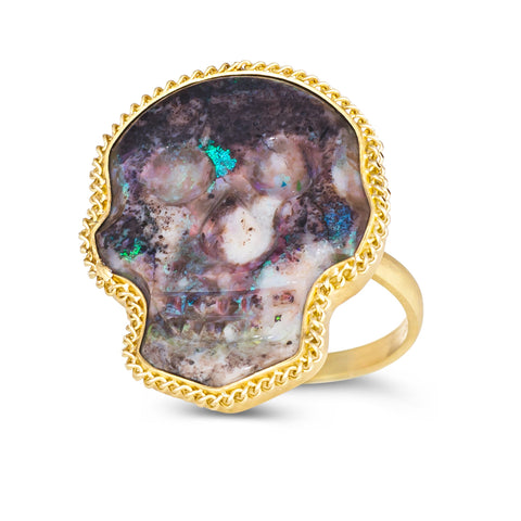 A large purple hued opal is carved into a skull and set in a braided 18k yellow gold bezel. The stone rests on a thin ring band.