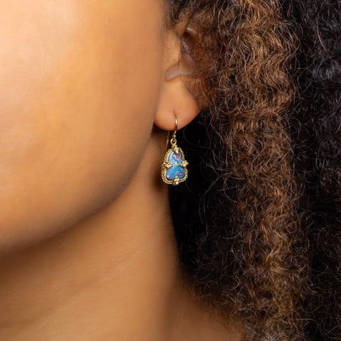 A model wears a small Boulder opal earring set in an 18k yellow gold chain wrapped bezel with four beaded prongs. The stone hangs on a French hook closure.