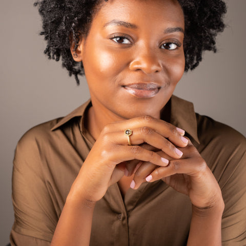 A model is wearing a small hexagon shaped charcoal colored diamond ring set in 18k yellow gold.
