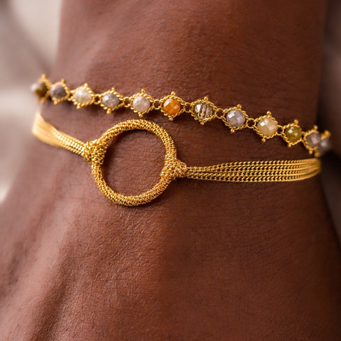 A model wears a stack of two bracelets, one featuring woven multi-colored diamond beads, and the other with a stardust effect circle in the center of three chains.