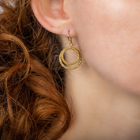 A model wears a small 18k yellow gold earring featuring two interlocking circles crafted with chain to create a stardust-like effect.