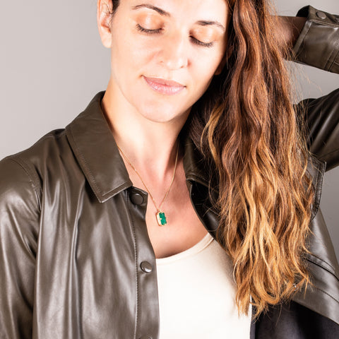 A model wears a rectangular emerald pendant set in an 18k yellow gold bezel wrapped in chain that hangs on a short delicate chain.
