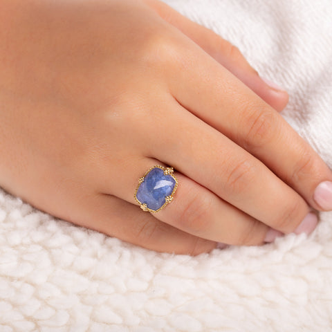 A model wears a large tanzanite stone ring is set in an 18k yellow gold chain wrapped bezel with four beaded prongs. The stone sits on a thin band.