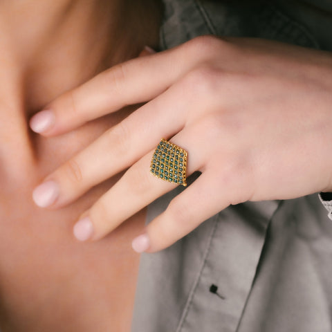 A model wears an 18k yellow gold ring crafted with blue diamond beads that are woven into a diamond shaped lattice pattern.