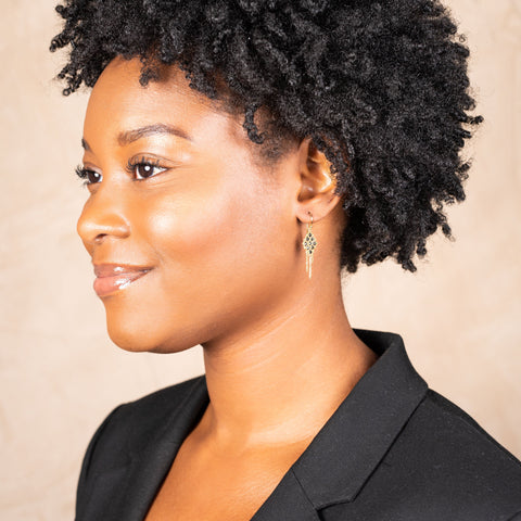 A model wears an 18k yellow gold earring crafted with black diamonds woven into a diamond shaped lattice pattern and has three dangling chains at the bottom.