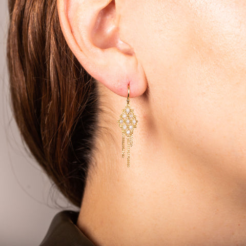 A model wears an 18k yellow gold earring crafted with silver diamonds woven into a diamond shaped lattice pattern and has three dangling chains at the bottom.