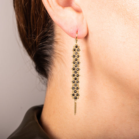 A pair of long black diamond earrings features a woven lattice pattern and two dangling chains at the bottom of the earring. The earrings fasten with french hook closures.