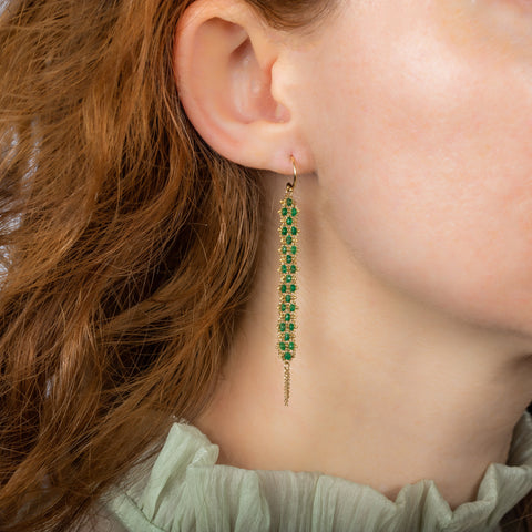 A pair of long green emerald earrings features a woven lattice pattern and two dangling chains at the bottom of the earring. The earrings fasten with french hook closures.