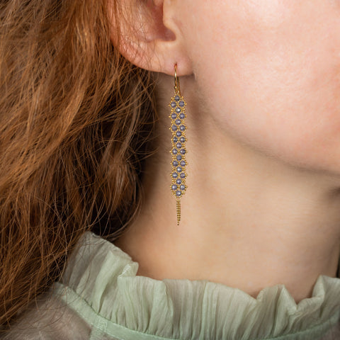 A model wears a long tanzanite earring featuring a woven lattice pattern and two dangling chains at the bottom of the earring. The earring fastens with french hook closures.