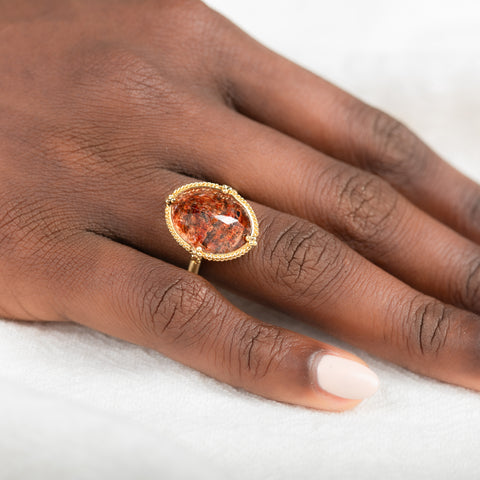 Gold ring featuring a sunstone gemstone on a person's finger against a neutral background