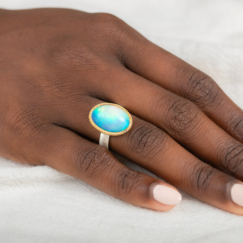 Hand wearing a ring with an Ethiopian opal gemstone set in a gold bezel with a silver band on a white background