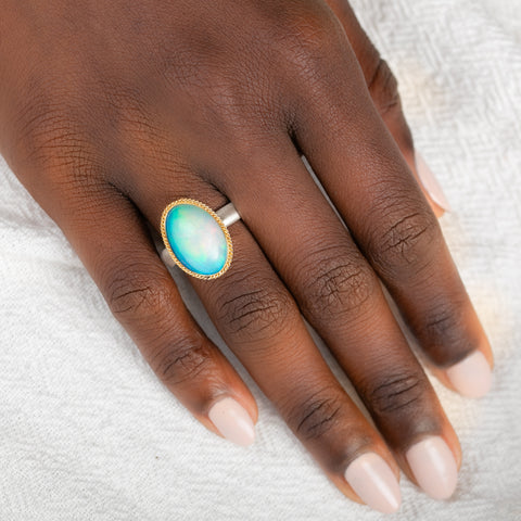 Hand wearing a ring with an Ethiopian opal gemstone set in a gold bezel with a silver band on a white background