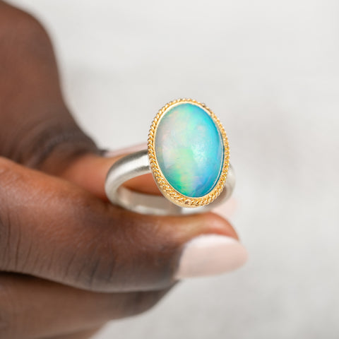 Hand holding a ring with an Ethioipian opal gemstone set in a gold bezel with a silver band on a white background