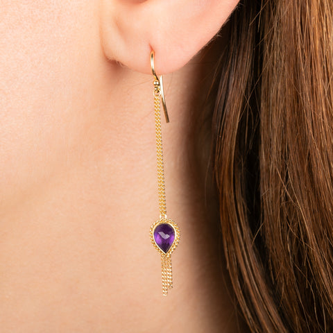 A gold dangle earring with a amethyst gemstone, shown worn on a person's ear.