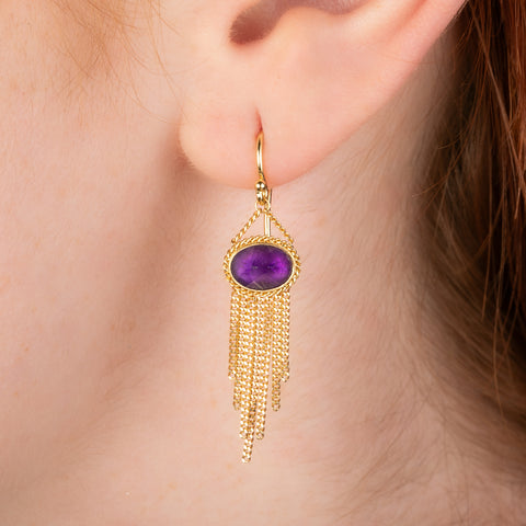 Gold earring featuring purple amethyst gemstone, shown worn in a person's ear.