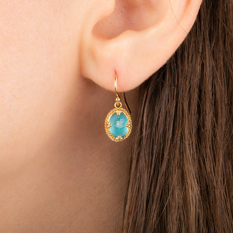 A close-up of a person's ear wearing a gold earring with an oval apatite gemstone in a textured gold bezel setting.