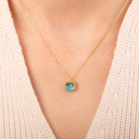 A gold chain necklace with a square pendant featuring an apatite stone set in an 18k bezel, worn on a person's neck.