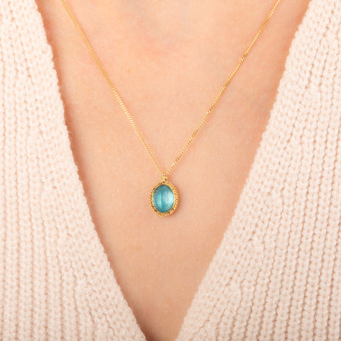 A gold chain necklace with an oval pendant featuring an apatite stone set in an 18k bezel, worn on a person's neck.
