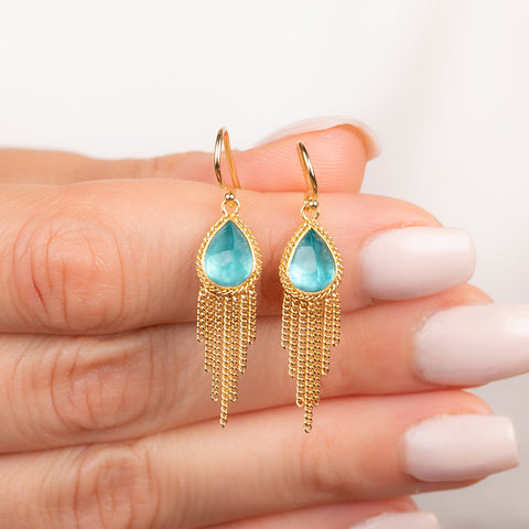 A pair of gold dangle earrings featuring teardrop-shaped apatite gemstones and cascading gold chain fringe, shown held on a person's finger