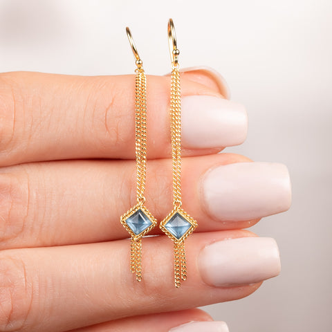 A pair of gold dangle earrings with textured chains leading to aquamarine gemstones, shown held in hand.
