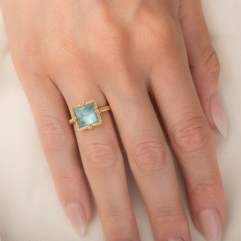 A square-cut light blue aquamarine ring set in an 18k gold bezel, worn on a person's finger.