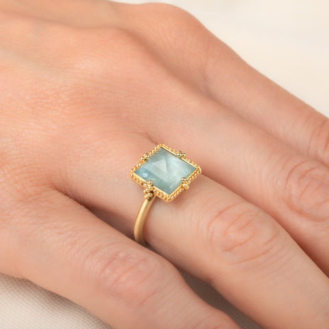 A square-cut light blue aquamarine ring set in an 18k gold bezel, worn on a person's finger.