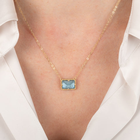Gold necklace with an aquamarine gemstone pendant worn by a person in a white shirt.
