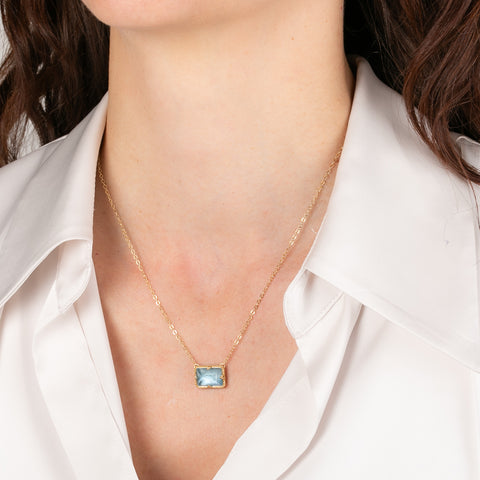 Gold necklace with an aquamarine gemstone pendant worn by a person in a white shirt.